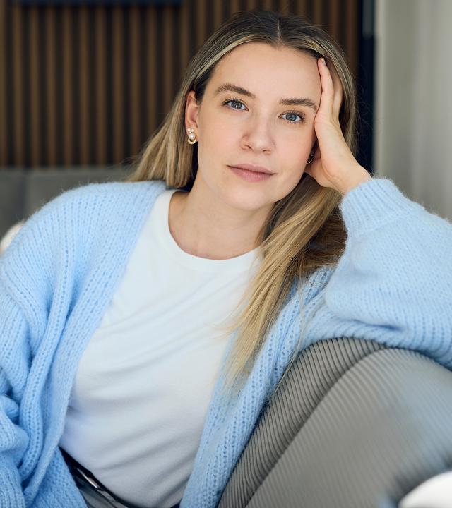 37°Leben: Deutschland, wann bin ich genug?": Valentina, eine blonde junge Frau sitzt auf einem grauen Sofa in einer modernen Wohnung. Sie stützt ihren Kopf mit der Hand ab und blickt ruhig und nachdenklich in die Kamera.