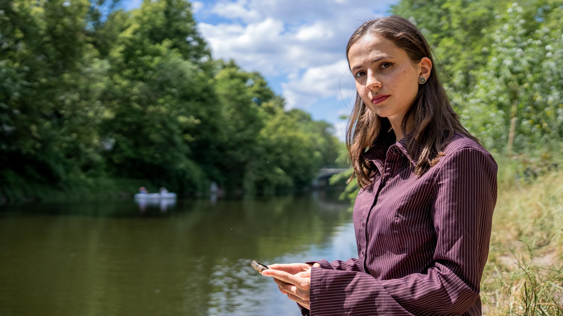 "37°Leben: Deepfakes - Leben nach digitaler Gewalt": Frau mit offenen braunen Haaren und gestreifter Bluse steht am Ufer eines Flusses und schaut angespannt. In ihren Händen hält sie ein Handy.