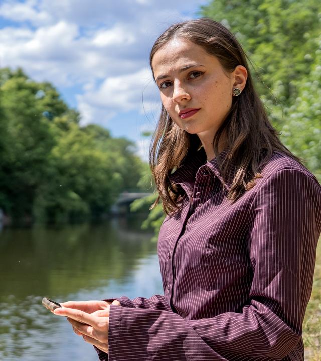 "37°Leben: Deepfakes - Leben nach digitaler Gewalt": Frau mit offenen braunen Haaren und gestreifter Bluse steht am Ufer eines Flusses und schaut angespannt. In ihren Händen hält sie ein Handy.