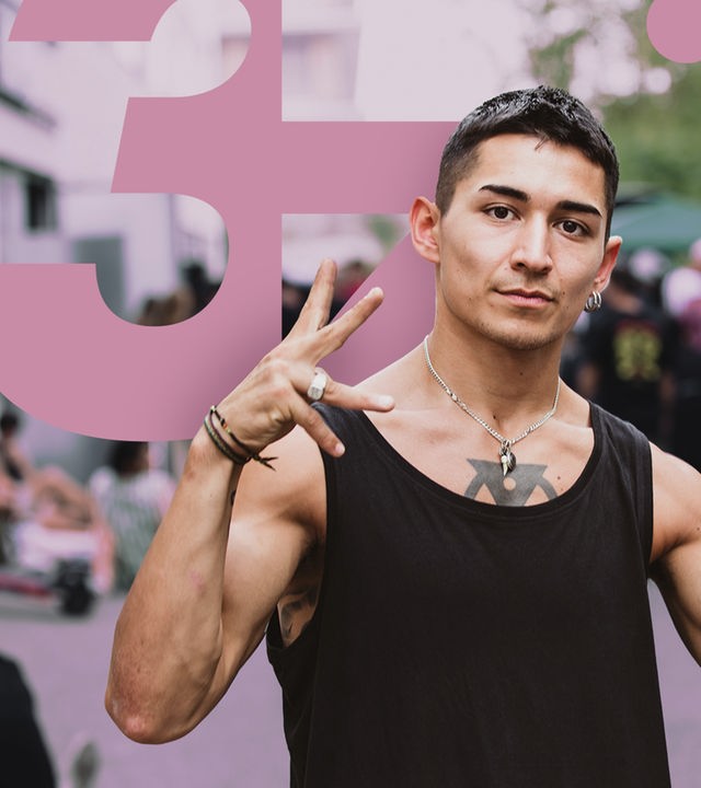 Ein junger Mann posiert auf der Straße, im Hintergrund, verschwommen zu erkennen, stehen einige Menschen beisammen