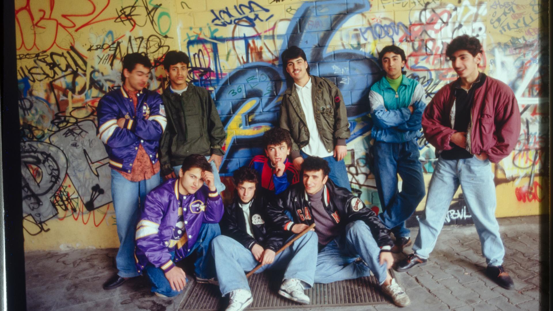 Foto einer Gruppe männlicher Jugendlicher in 1980er Jahre Klamotten vor einer mit Graffitti besprühten gelb gekachelten Wand.