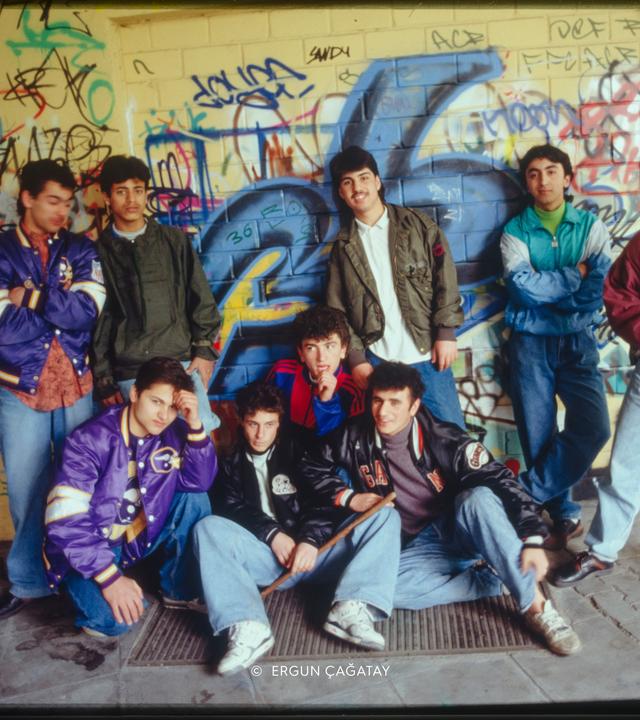 Foto einer Gruppe männlicher Jugendlicher in 1980er Jahre Klamotten vor einer mit Graffitti besprühten gelb gekachelten Wand.
