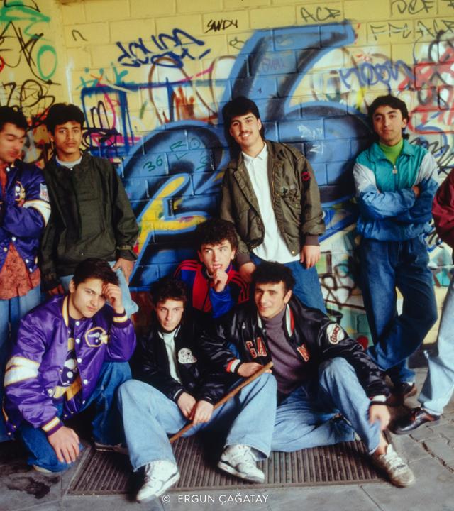 Foto einer Gruppe männlicher Jugendlicher in 1980er Jahre Klamotten vor einer mit Graffitti besprühten gelb gekachelten Wand.