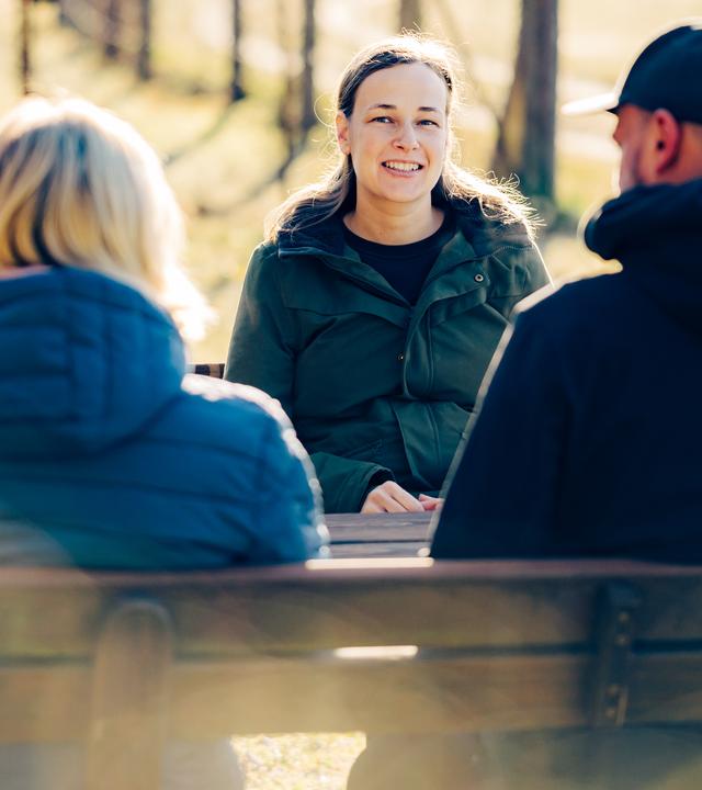Elisa steht an ein Ortsschild aus Holz gelehnt und lächelt in die Kamera. Auf dem Schild steht "Willkommen in Quarnebeck" neben einem Wappen.