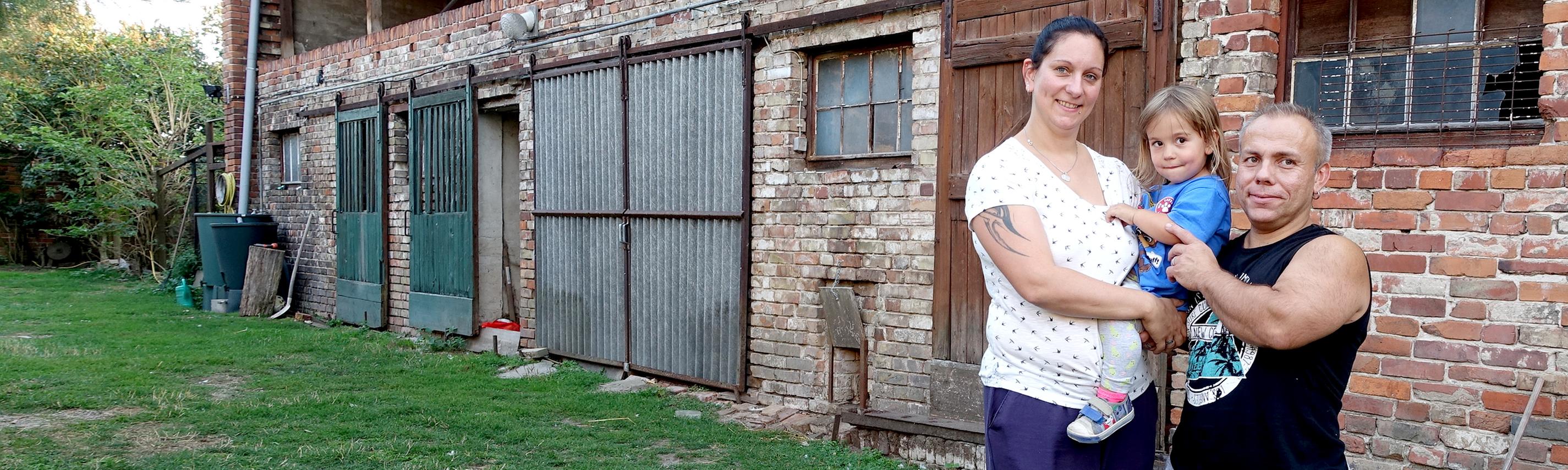 Ralf und seine Frau Susi stehen mit ihrer kleinen Tochter vor ihrem Haus. Susi ist einen Kopf größer als ihr Mann.