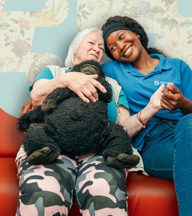 Zwei Frauen sitzen auf einem roten Sofa, die jüngere hat liebevoll den Arm um die ältere Frau gelegt.