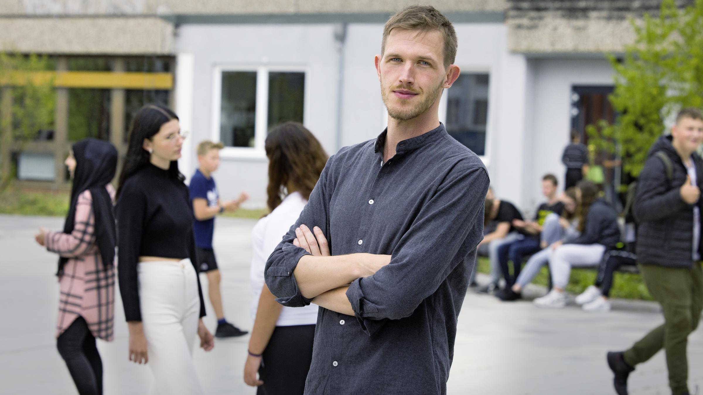 "37°: Kaltstart im Klassenzimmer - Quereinsteiger als Lehrer": Till (35) steht mit verschränkten Armen auf dem Schulhof. Um ihn herum sind Schüler und Schülerinnen in der Hofpause.