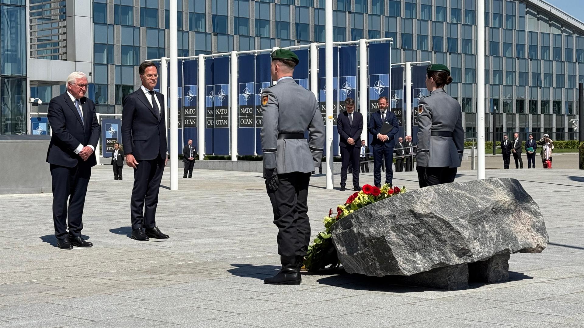 Steinmeier und Rutte bei Festakt in Brüssel. Militärangehörige stehen ihnen gegenüber, zwischen ihnen ein Blumenkranz