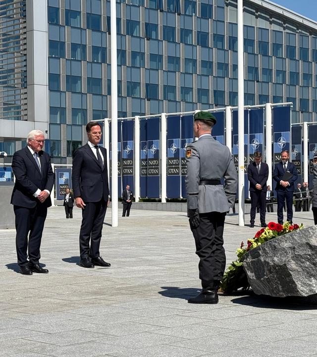 Steinmeier und Rutte bei Festakt in Brüssel. Militärangehörige stehen ihnen gegenüber, zwischen ihnen ein Blumenkranz
