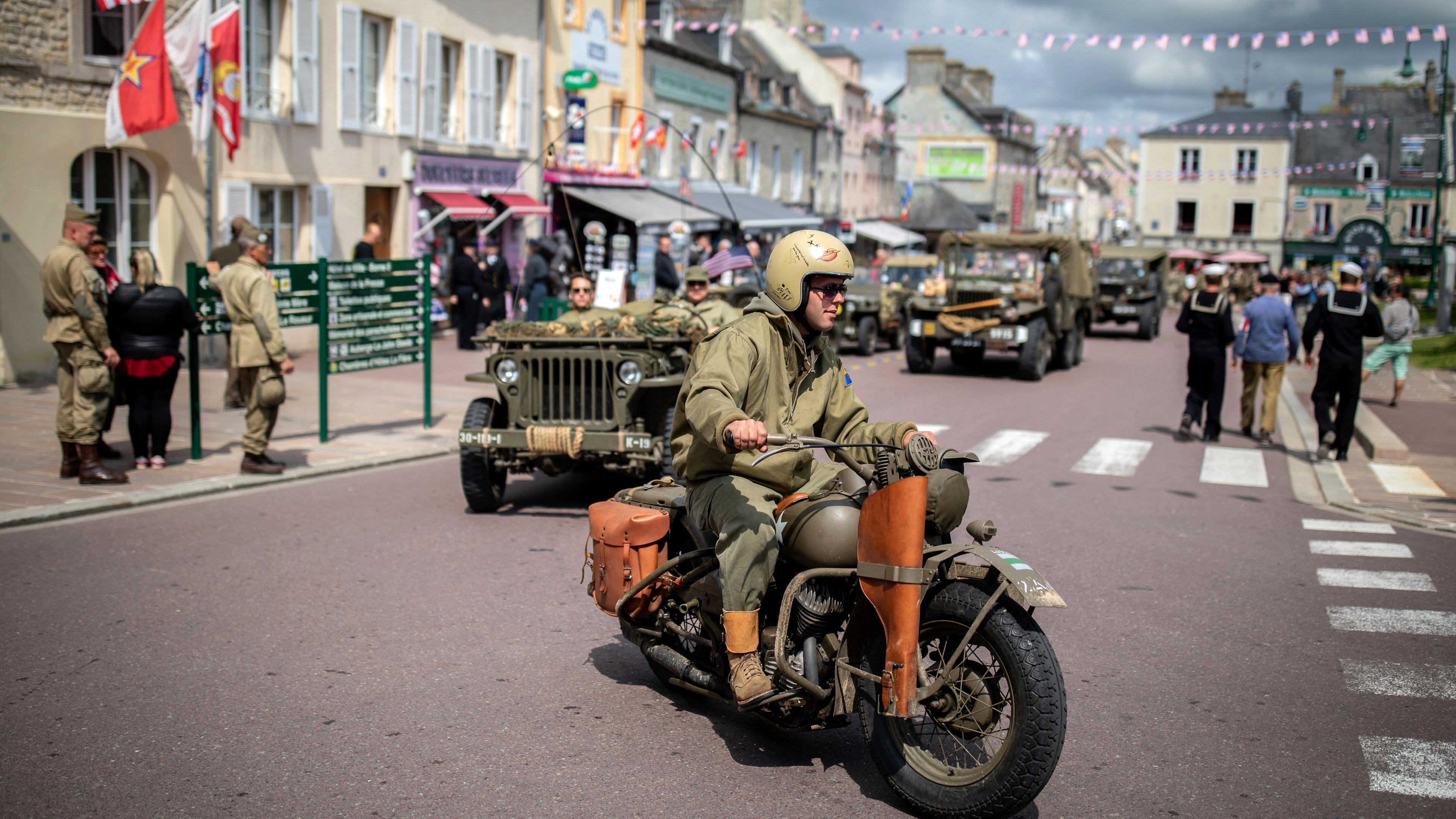 In historischen US-Uniformen fahren Menschen auf ihren Fahrzeugen aus dem Zweiten Weltkrieg durch den Ort Sainte Mere Eglise (Frankreich) am 03.06.2019