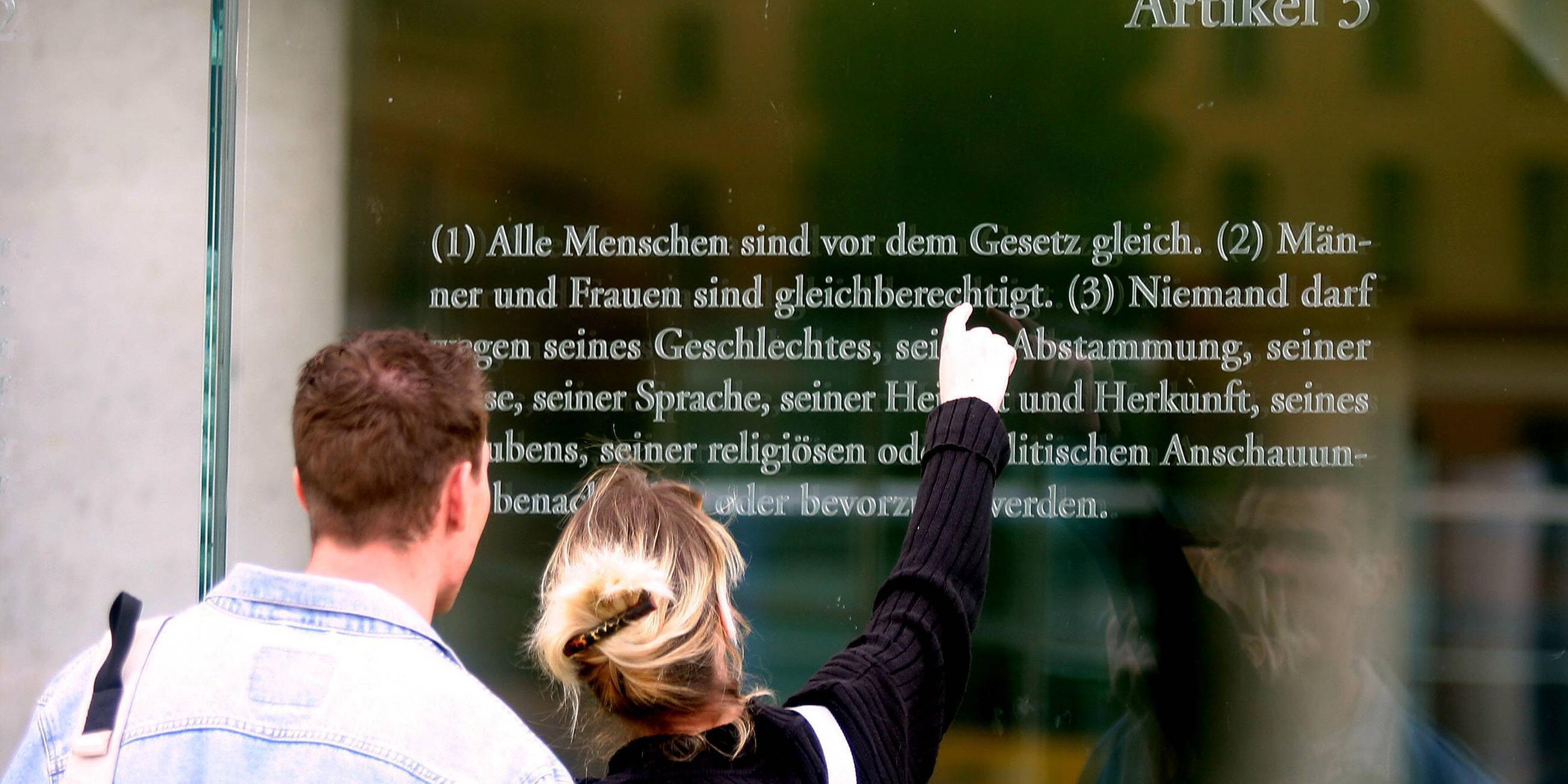 Ein Mann und eine Frau stehen vor einer Glaswand, auf der Artikel 3 des Grundgesetzes zu lesen ist.