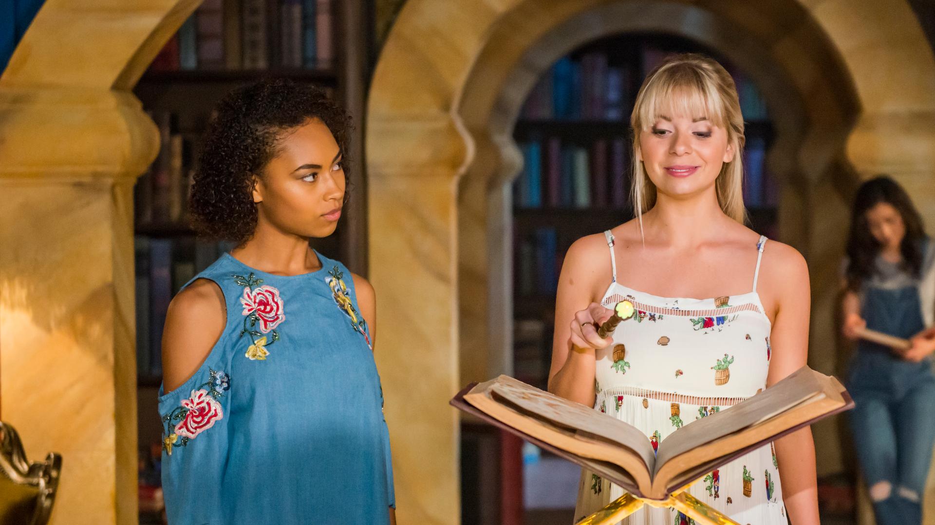 Lily steht in der Bibliothek vor einem dicken Zauberbuch mit ihrem Zauberstab in der Hand.