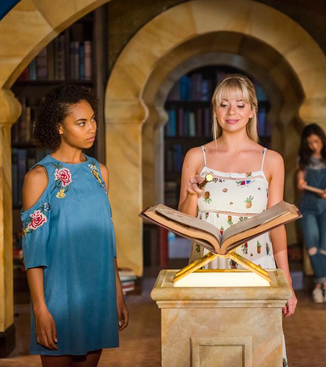 Lily steht in der Bibliothek vor einem dicken Zauberbuch mit ihrem Zauberstab in der Hand.