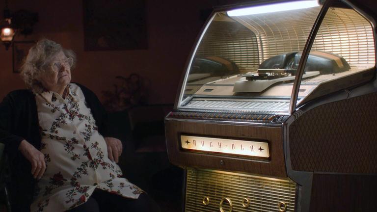 "80.000 Schnitzel": Eine ältere Dame mit grauen Haaren und einer Hornbrille, Berta Zenefels, sitzt in einer abgedunkelten Gastwirtschaft vor einer beleuchteten Musikbox aus den 50er Jahren. Sie hört der Musik zu, die in der Musikbox von einer Schallplatte abgespielt wird.