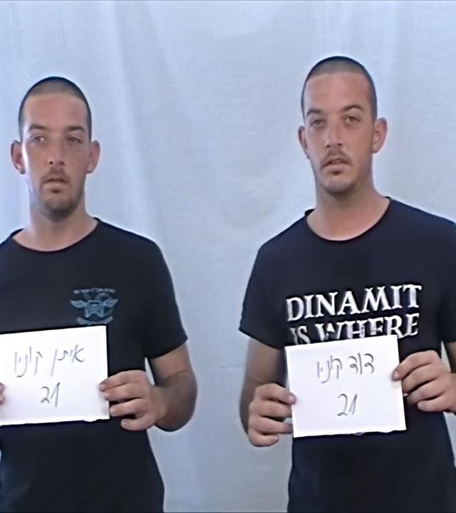 Zwei gleich aussehende Zwillings-Männer in T-Shirts stehen nebeneinander und halten Zettel mit Nummern hoch.