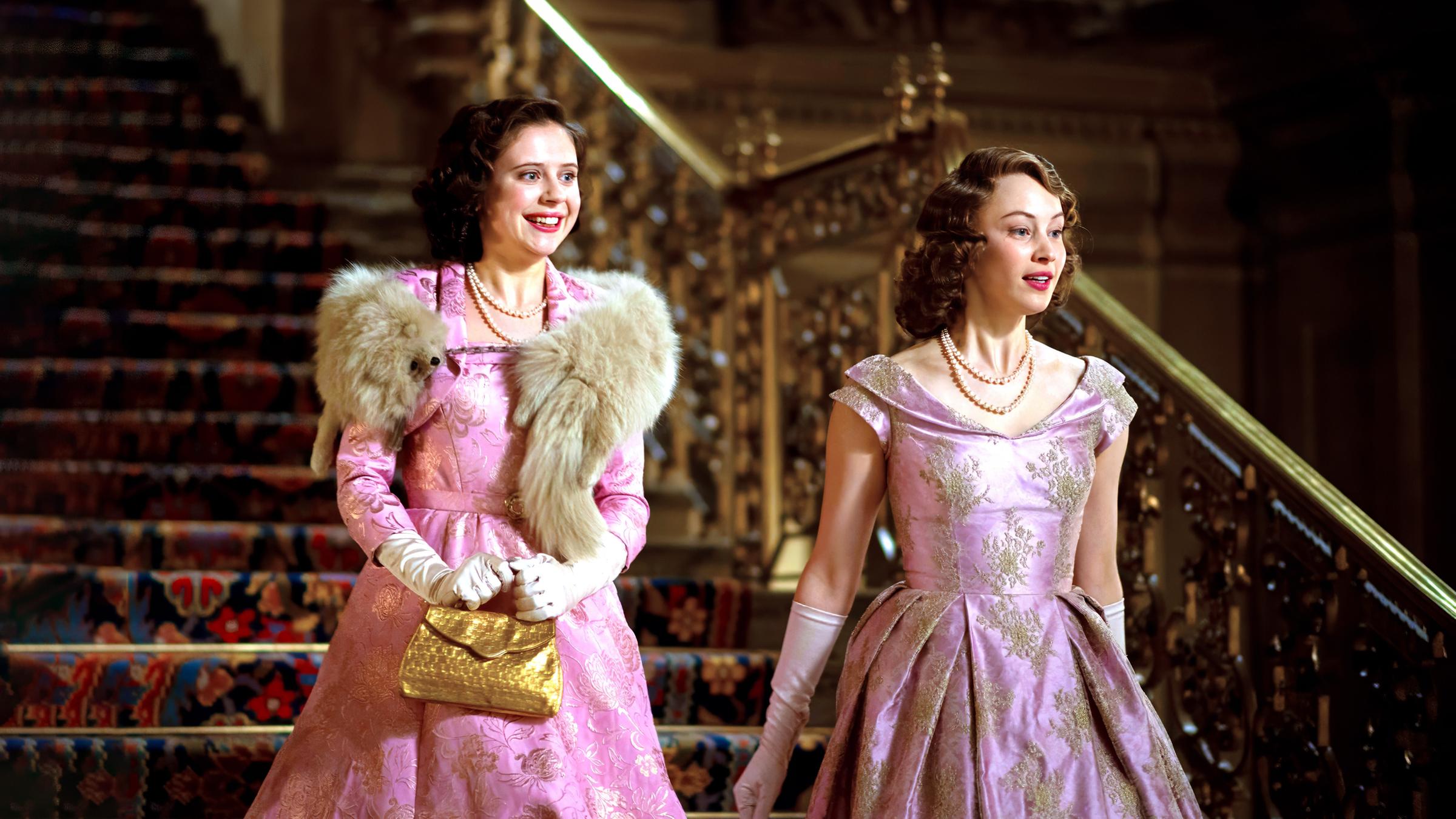 Die beiden Prinzessinnen Margaret (Bel Powley) und Elizabeth (Sarah Gadon) kommen nebeneinander eine prunkvolle Treppe herunter. Sie schauen sich lächelnd um. Beide tragen fast identische, elegante Kleider.