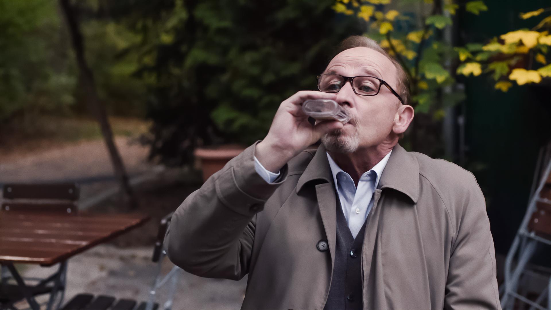 Ludwig Schaller (Alexander Held) sitzt im Grünen in einem Café und trinkt Hochprozentigen aus einem Fläschchen. 
