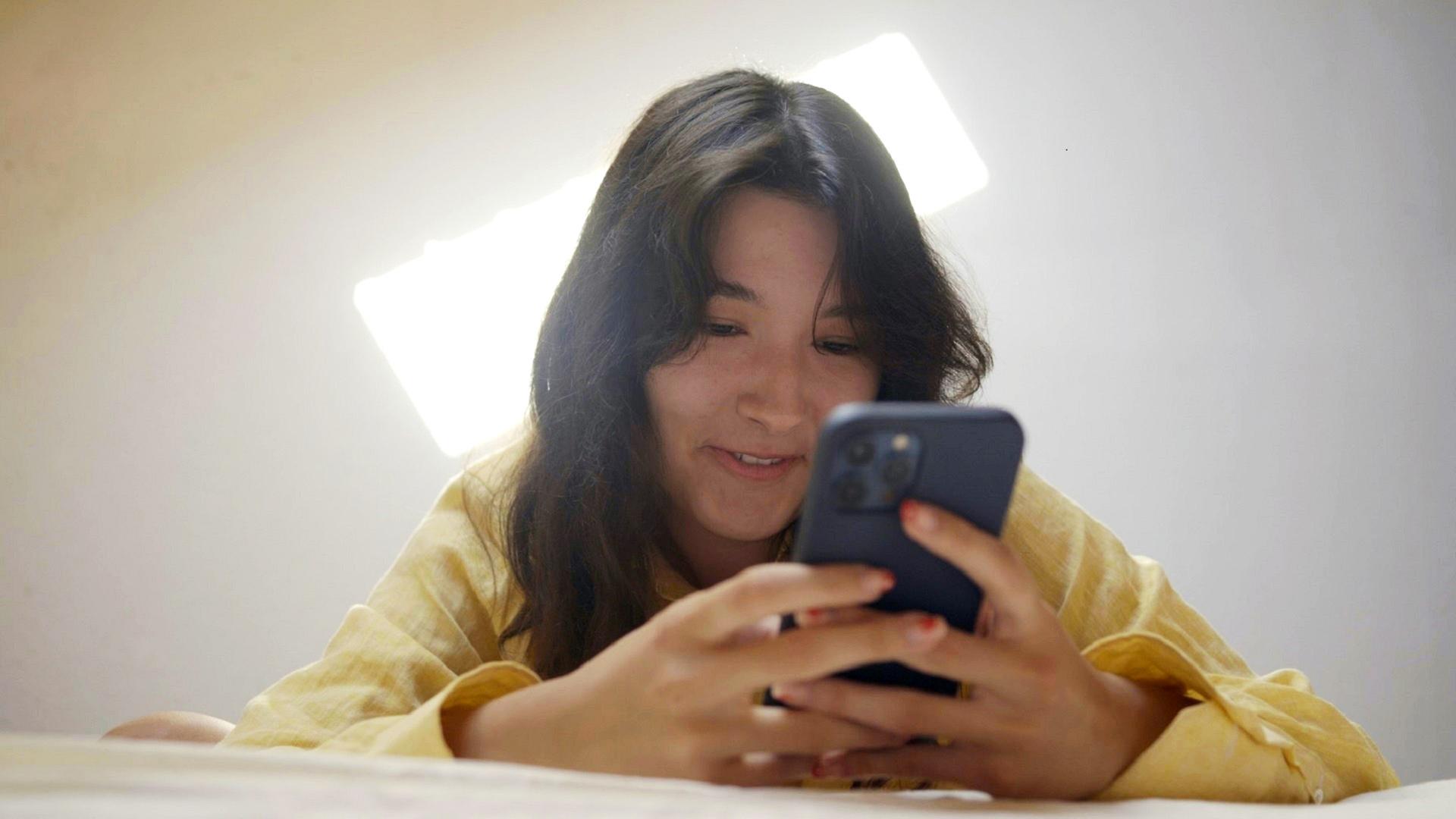 Eine junge Frau mit langen braunen Haaren und einer gelben langärmeligen Bluse liegt bäuchlings auf einem Bett und betrachtet lächelnd etwas auf ihrem Smartphone. Über ihr erhellt eine Lampe mit weißem Licht den Raum.