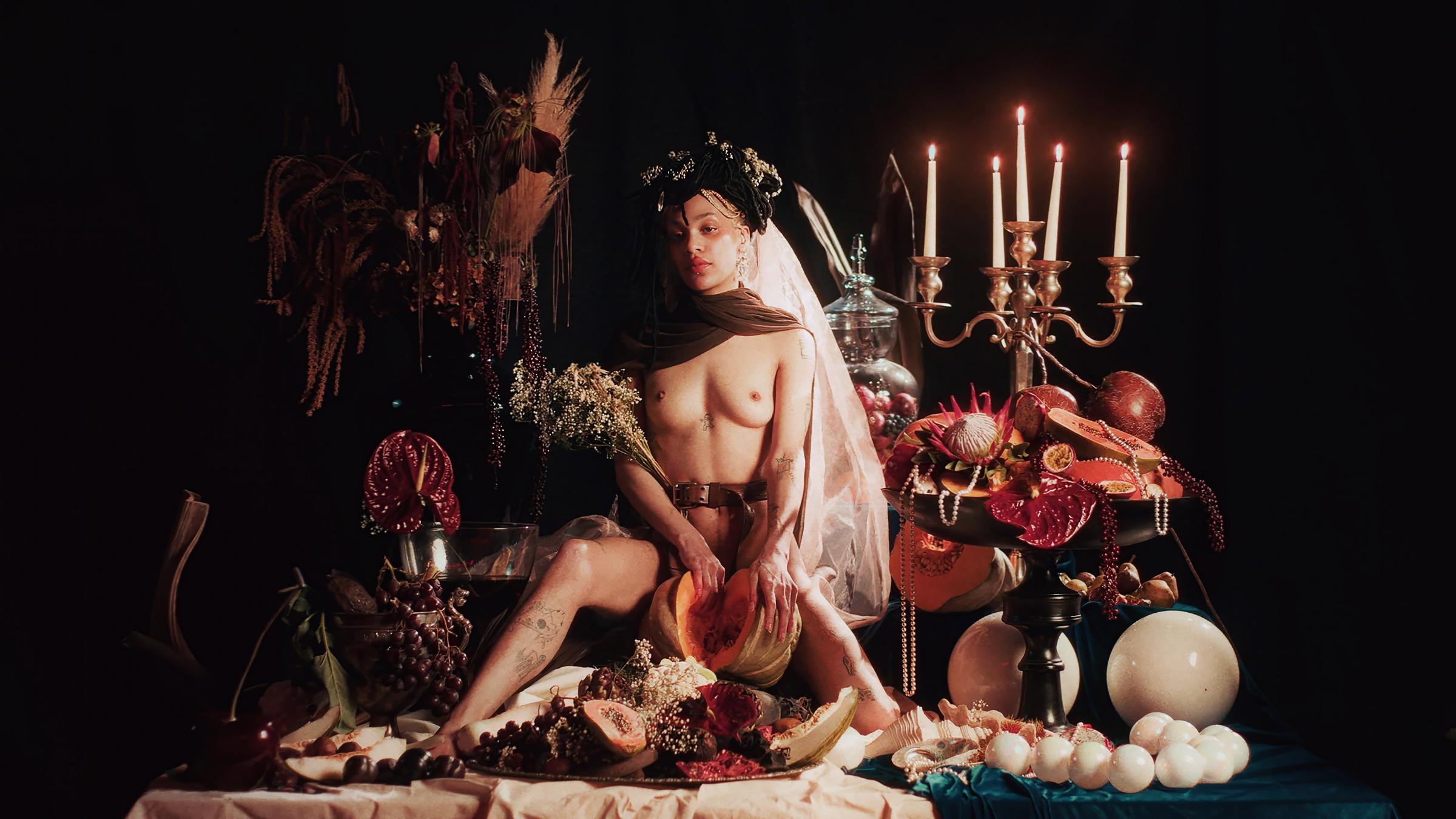 Eine nackte junge Frau mit Blumenschmuck im Haar sitzt auf einem opulent arrangierten Tisch voller Früchte und Blumen.