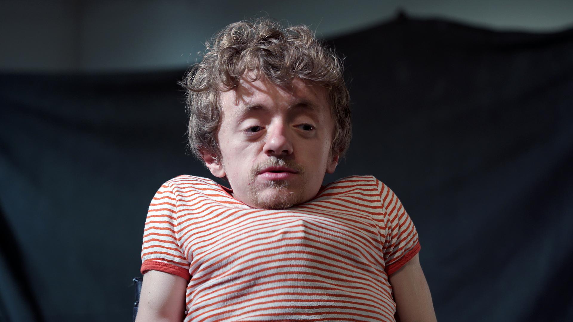Leonard, ein kleinwüchsiger junger Mann, trägt ein rotgestreiftes T-Shirt.
