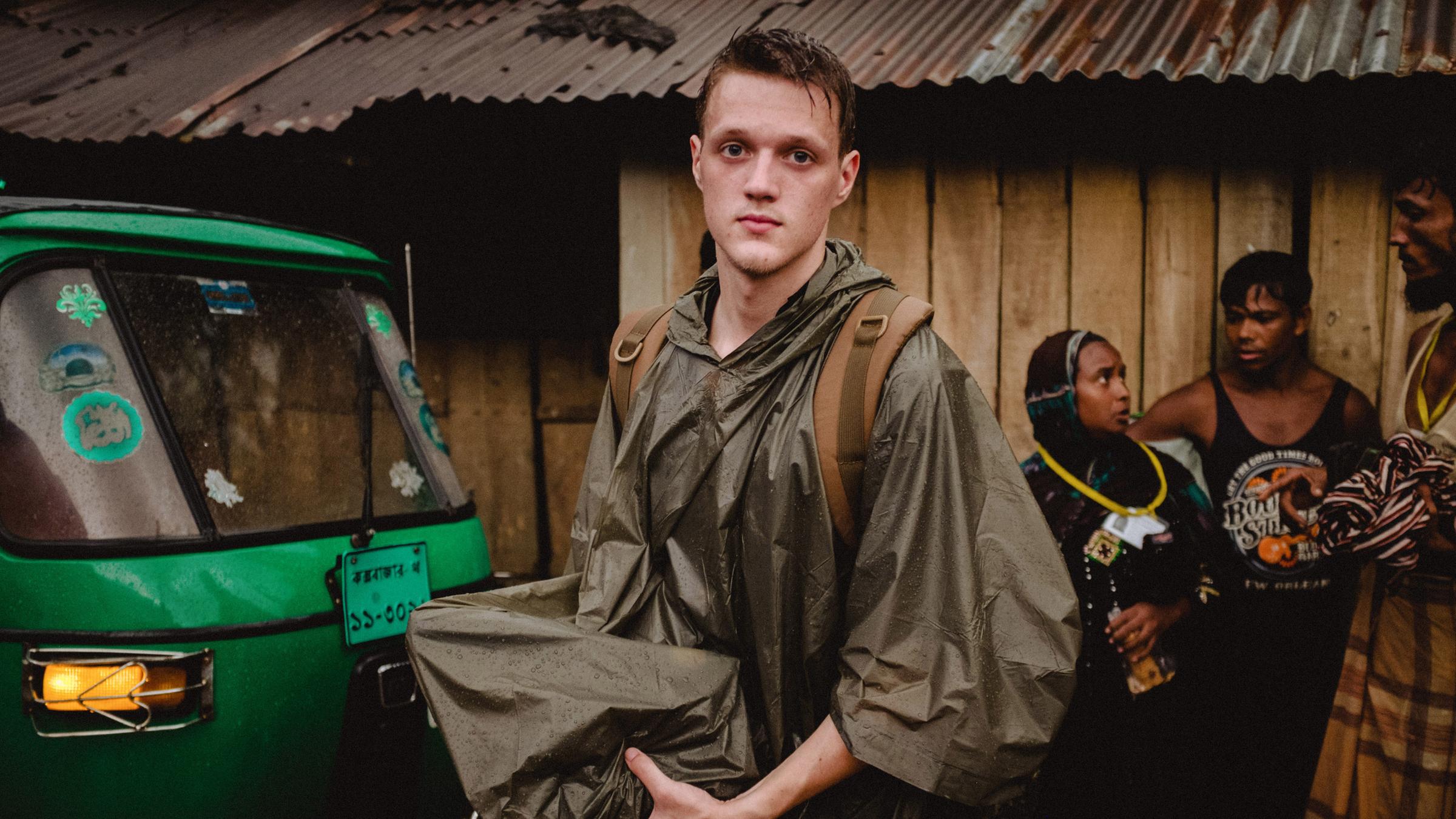 "Ab 18! – Hinter unserem Horizont": Halbnahe Aufnahme eines jungem Mannes, der unter einem Regenponcho seine Kamera schützt und in die Kamera blickt.