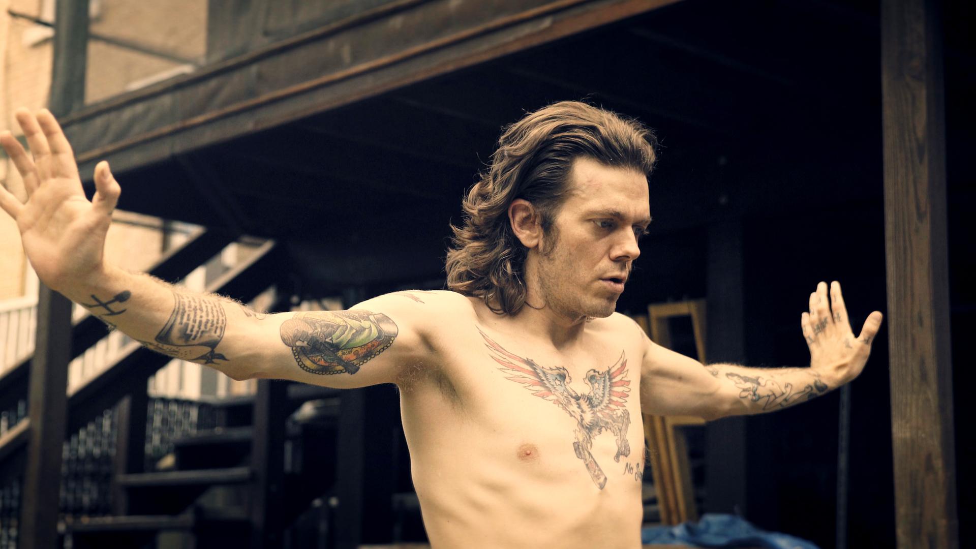 Ab 18! - Joe Boots - Tätowierter Mann mit ausgebreiteten Armen.