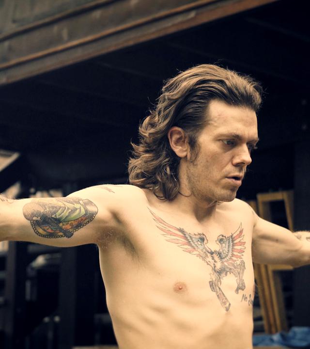 Ab 18! - Joe Boots - Tätowierter Mann mit ausgebreiteten Armen.