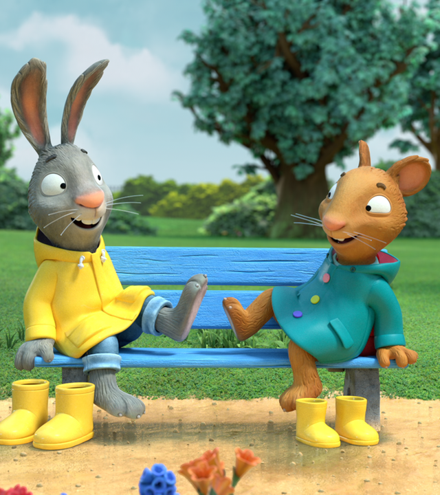 Pip und Posy sitzen in ihren Regenmänteln auf einer Bank und klatschen sich mit ihren Füssen lachend ab. Vor der Bank stehen jeweils ein Paar gelbe Gummistiefel.