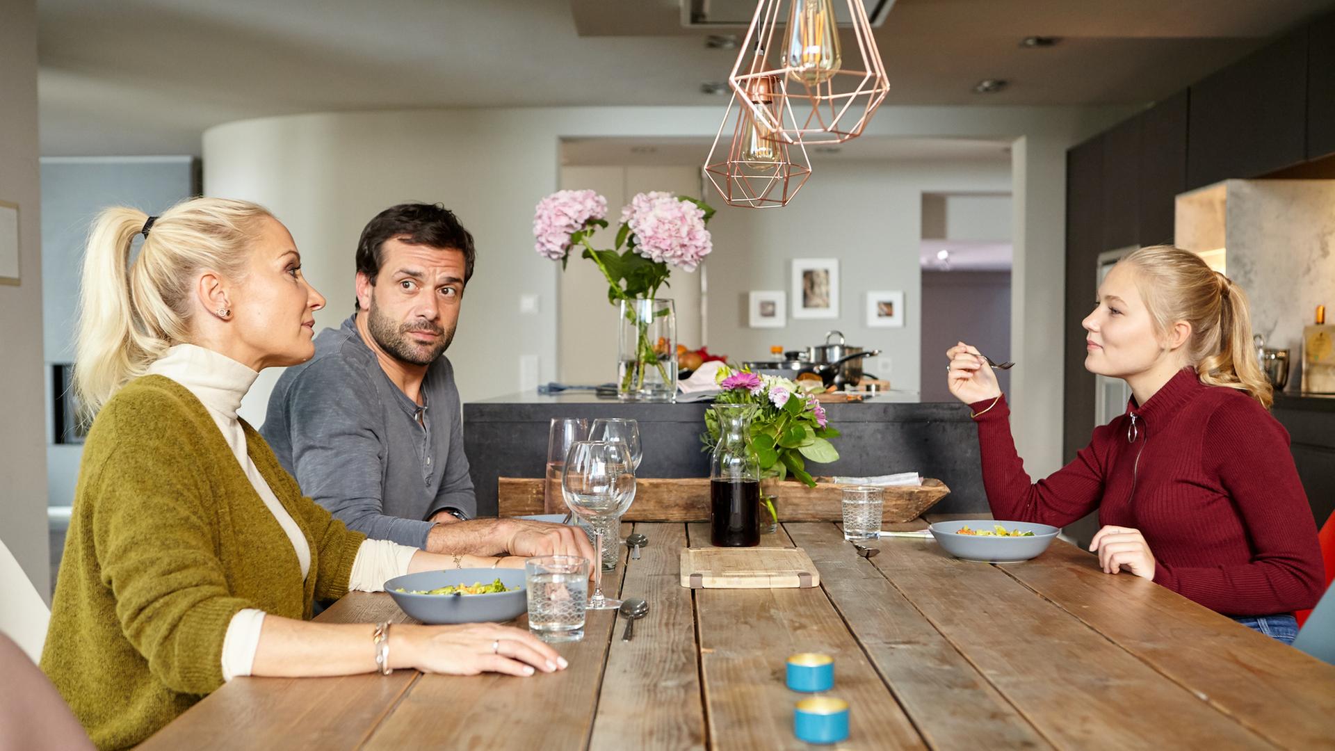 "Heldt - Ab in die Tonne": Ellen Bannenberg (Janine Kunze) und Nikolas Heldt (Kai Schumann) sitzen an einem Esstisch, auf dem Essgeschirr, Blumen und Kerzen stehen. Ihnen gegenüber sitzt Ellens Tochter Emily (Lili Budach), die eine Gabel in der Hand hält und die beiden lächelnd ansieht.