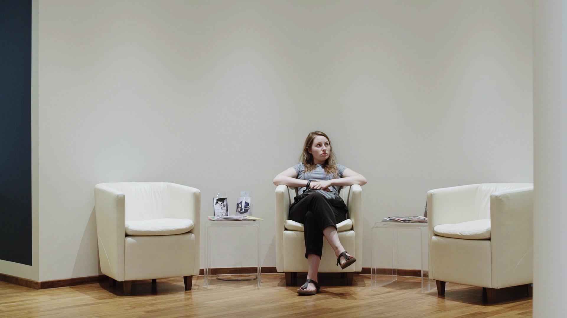 "Ab 18! – Dazwischen Elsa": Eine Sitzgruppe von drei Sesseln in einem Wartebereich. Die Protagonistin Elsa sitzt auf dem mittleren Sitz und blickt nach oben.