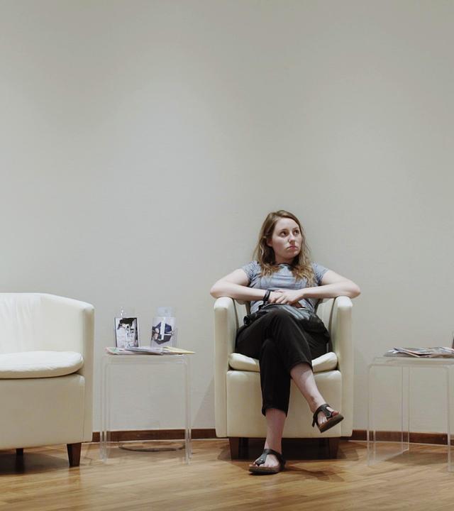 "Ab 18! – Dazwischen Elsa": Eine Sitzgruppe von drei Sesseln in einem Wartebereich. Die Protagonistin Elsa sitzt auf dem mittleren Sitz und blickt nach oben.