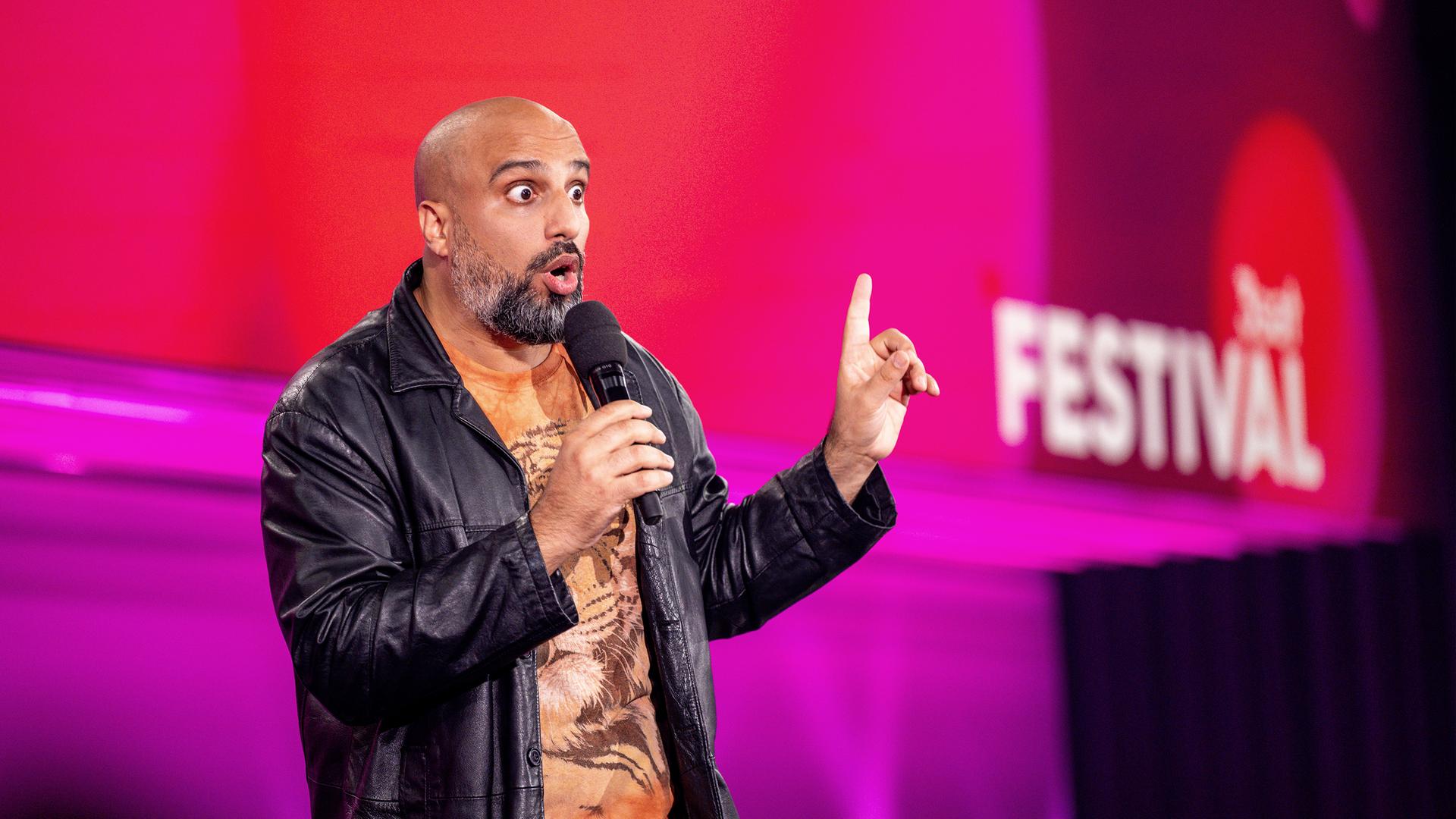 Abdelkarim steht in der Mitte des Bildes. In der rechten Hand hat er ein Mikrofon. Die linke Hand zeigt mit dem Zeigefinger nach oben. Der Hintergrund ist rot und lila.