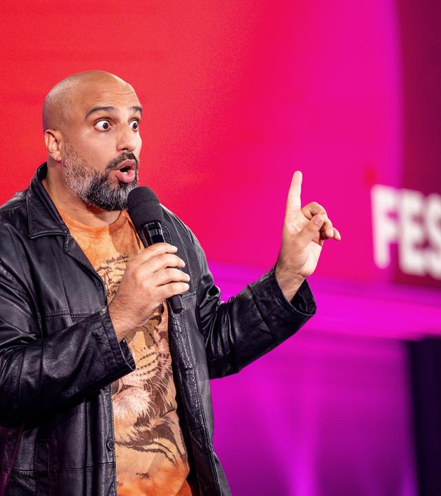 Abdelkarim steht in der Mitte des Bildes. In der rechten Hand hat er ein Mikrofon. Die linke Hand zeigt mit dem Zeigefinger nach oben. Der Hintergrund ist rot und lila.