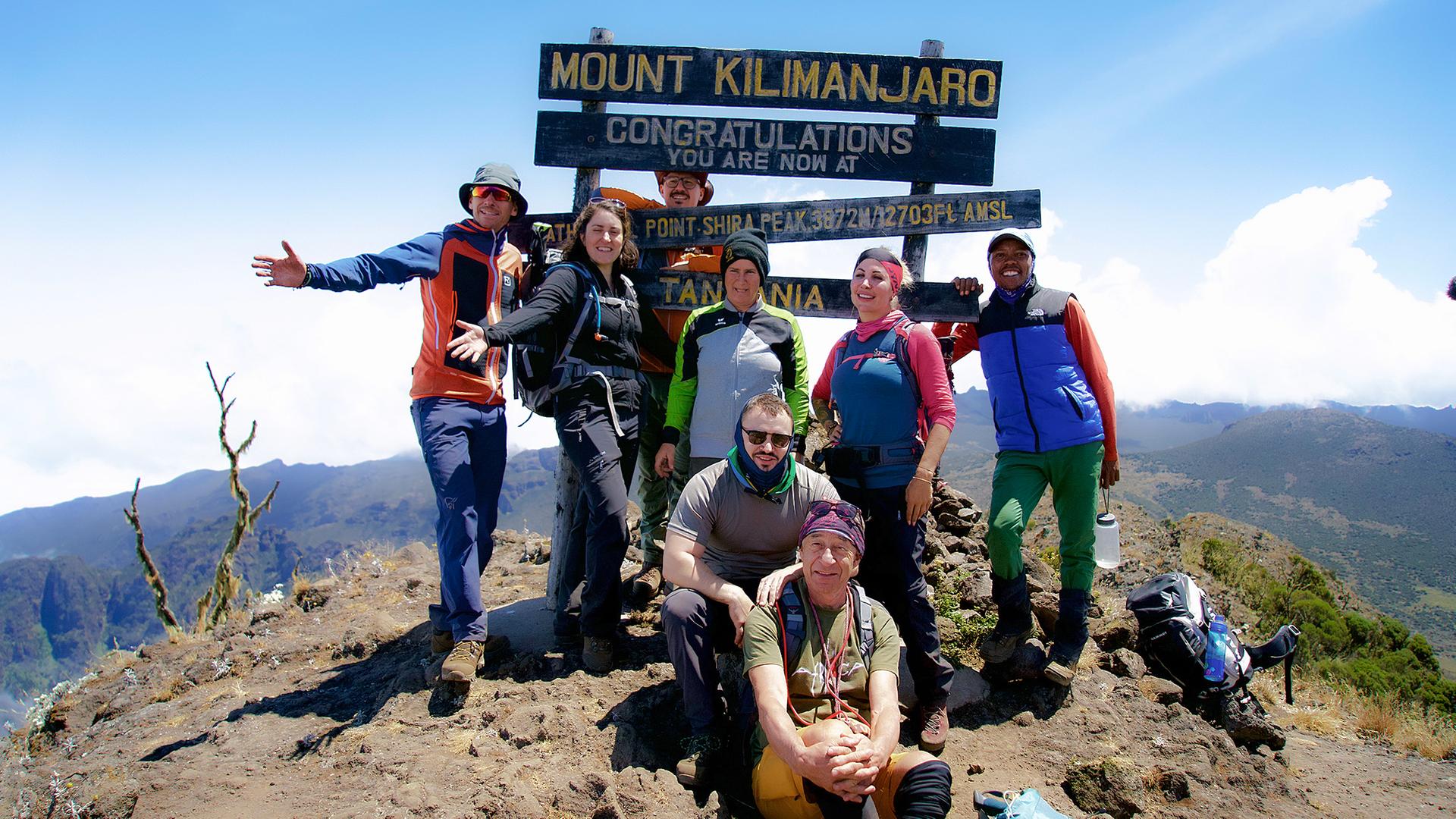 Acht Personen in Trekkingausrüstung posieren am Gipfel des Kilimandscharo für ein Gruppenfoto