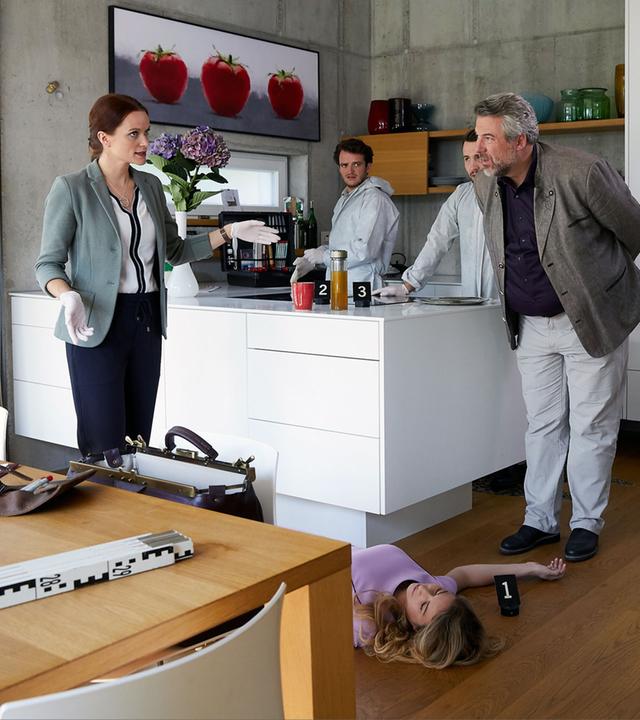 "Die Rosenheim-Cops - Abgerechnet wird zum Schluss": Die Kollegen Sandra Mai (Sina Wilke), Alois Kammermayr (Tommy Schwimmer), KTU-Kollege (Komparse), Anton Stadler (Dieter Fischer) und Christian Bach (Patrick Kalupa)der Kripo Rosenheim stehen in der Küche eines Hauses um eine Frau herum, die am Boden liegt.