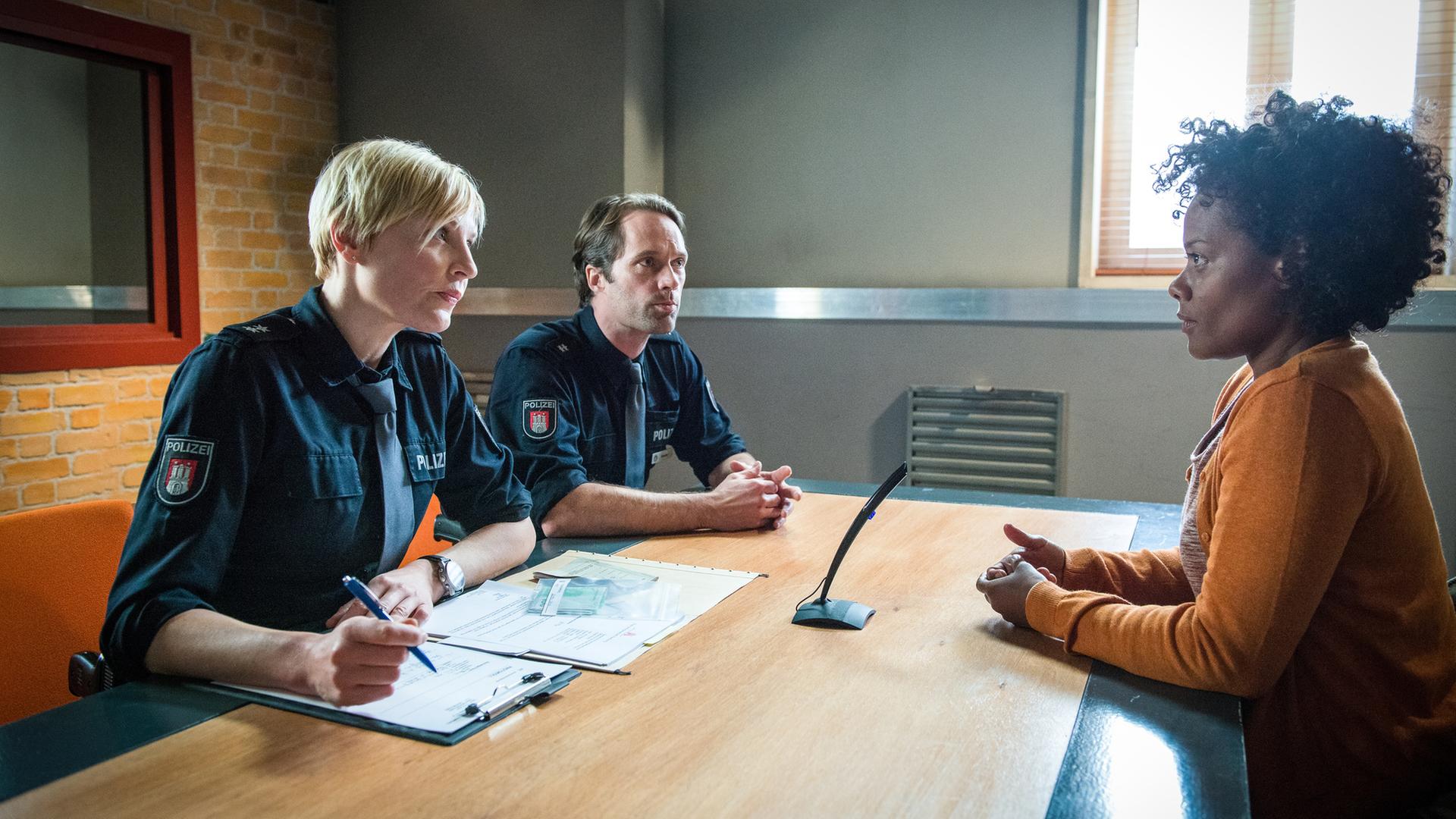 "Notruf Hafenkante - Abgeschoben": Im Befragungsraum sitzen die Polizeibeamten Melanie Hansen (Sanna Englund) und Mattes Seeler (Matthias Schloo) Ifemelu Okoye (Denise M'Baye) gegenüber. Melanie hat Unterlagen vor sich liegen und einen Stift in der Hand.