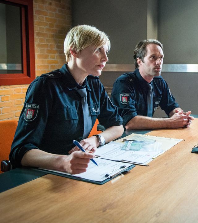 "Notruf Hafenkante - Abgeschoben": Im Befragungsraum sitzen die Polizeibeamten Melanie Hansen (Sanna Englund) und Mattes Seeler (Matthias Schloo) Ifemelu Okoye (Denise M'Baye) gegenüber. Melanie hat Unterlagen vor sich liegen und einen Stift in der Hand.