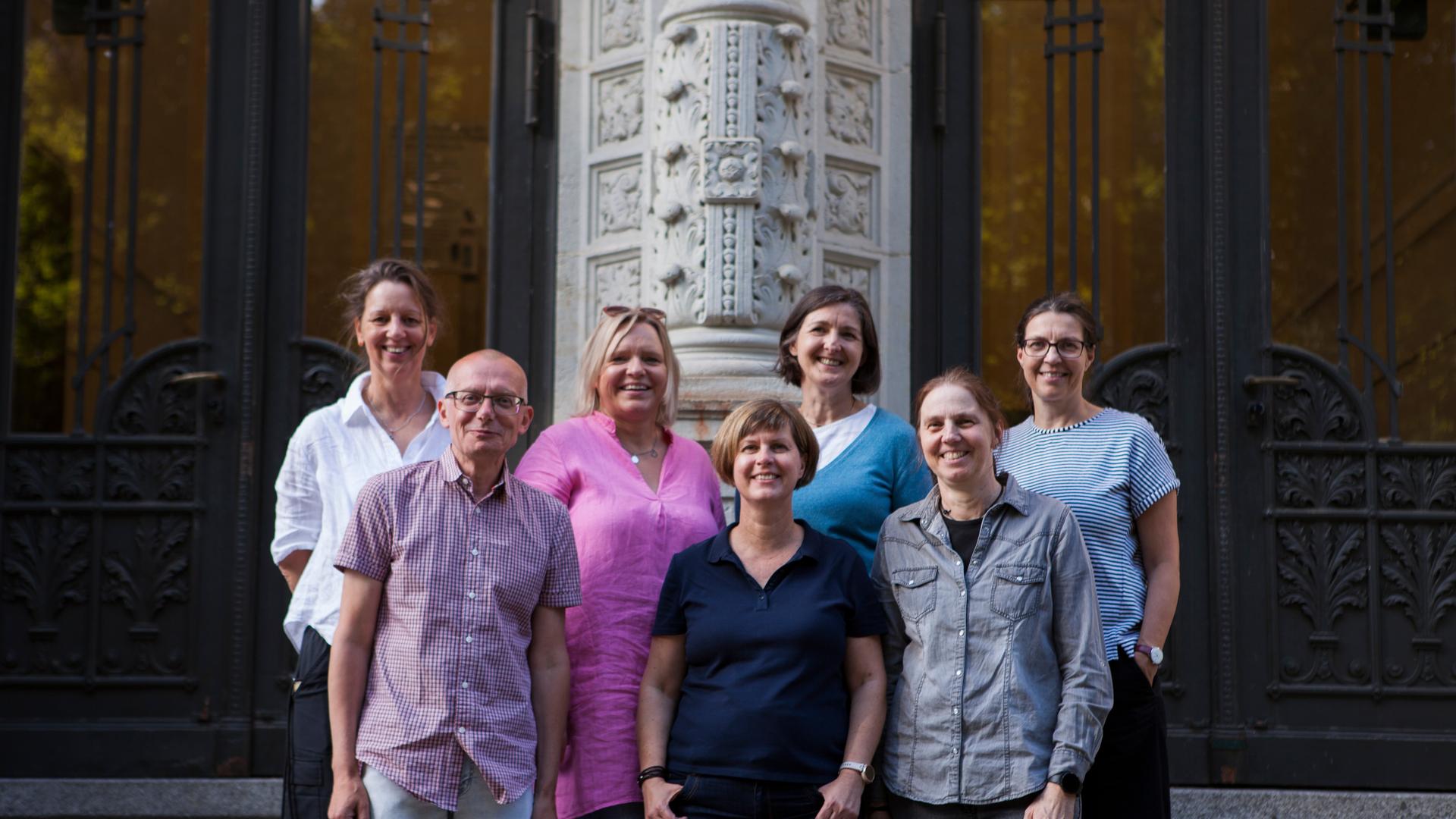 Sieben Erwachsene stehen vor einer Schule auf einer Treppe zum Foto verteilt.