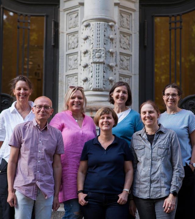 Sieben Erwachsene stehen vor einer Schule auf einer Treppe zum Foto verteilt.