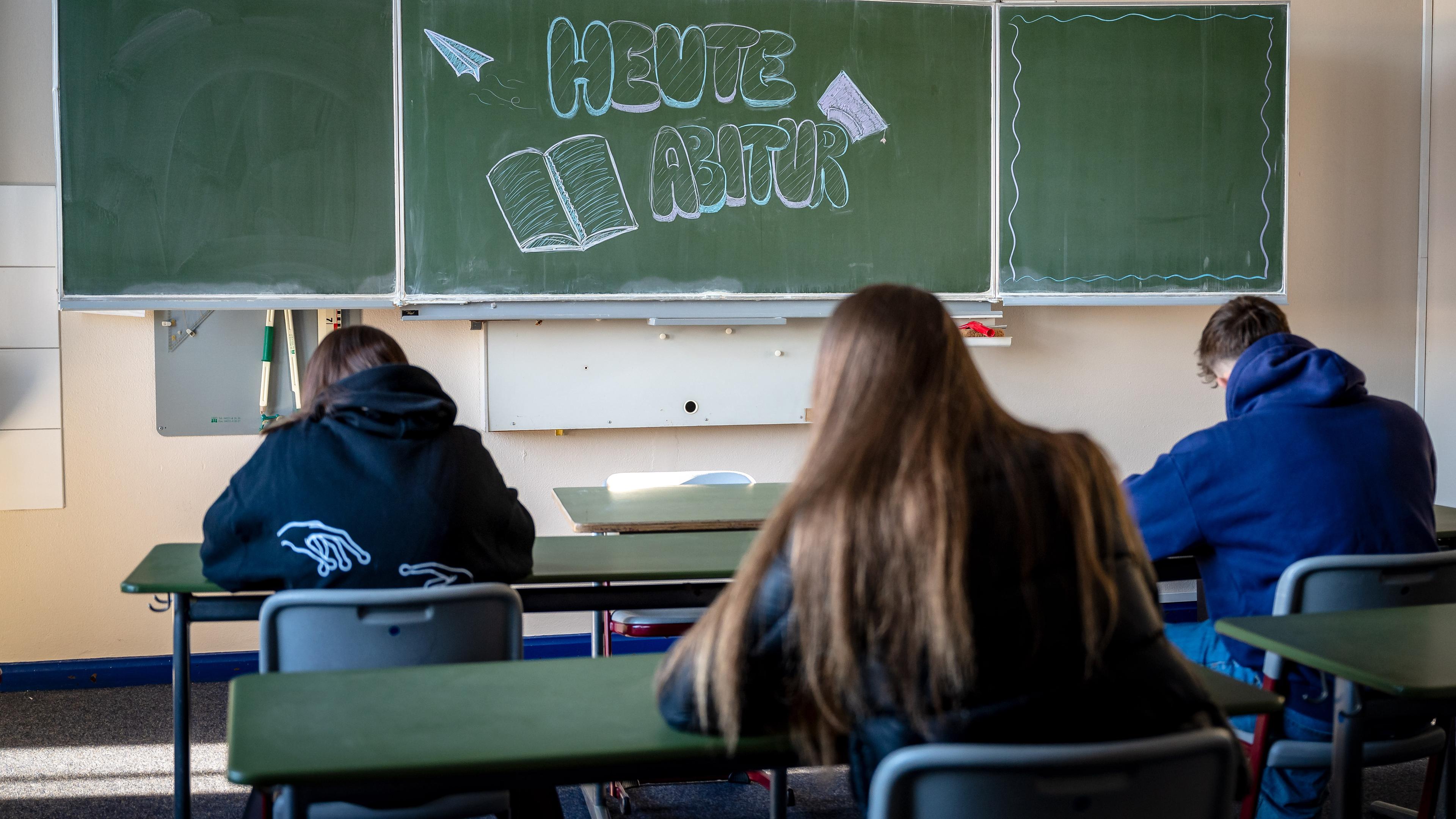 Ein Klassenzimmer. Drei Schüler und Schülerinnen sitzen an den Schultischen. An der Tafel steht: "Heute Abitur".