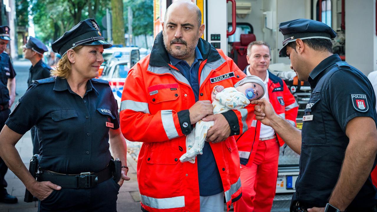 "Notruf Hafenkante - Abnabelung": Zwischen den Polizisten Claudia Fischer (Janette Rauch) und Tarik Coban (Serhat Cokgezen) steht der Arzt Dr. Aguilar (Gabriel Munoz Munoz). Er hält einen Säugling im Arm. Tarik streichelt das Gesicht des Säuglings, Claudia hat die Hände in die Hosentaschen gesteckt und schaut grinsend zu Tarik und dem Baby. Im Hintergrund der Notarztwagen und zwei Sanitäter (Komparsen).