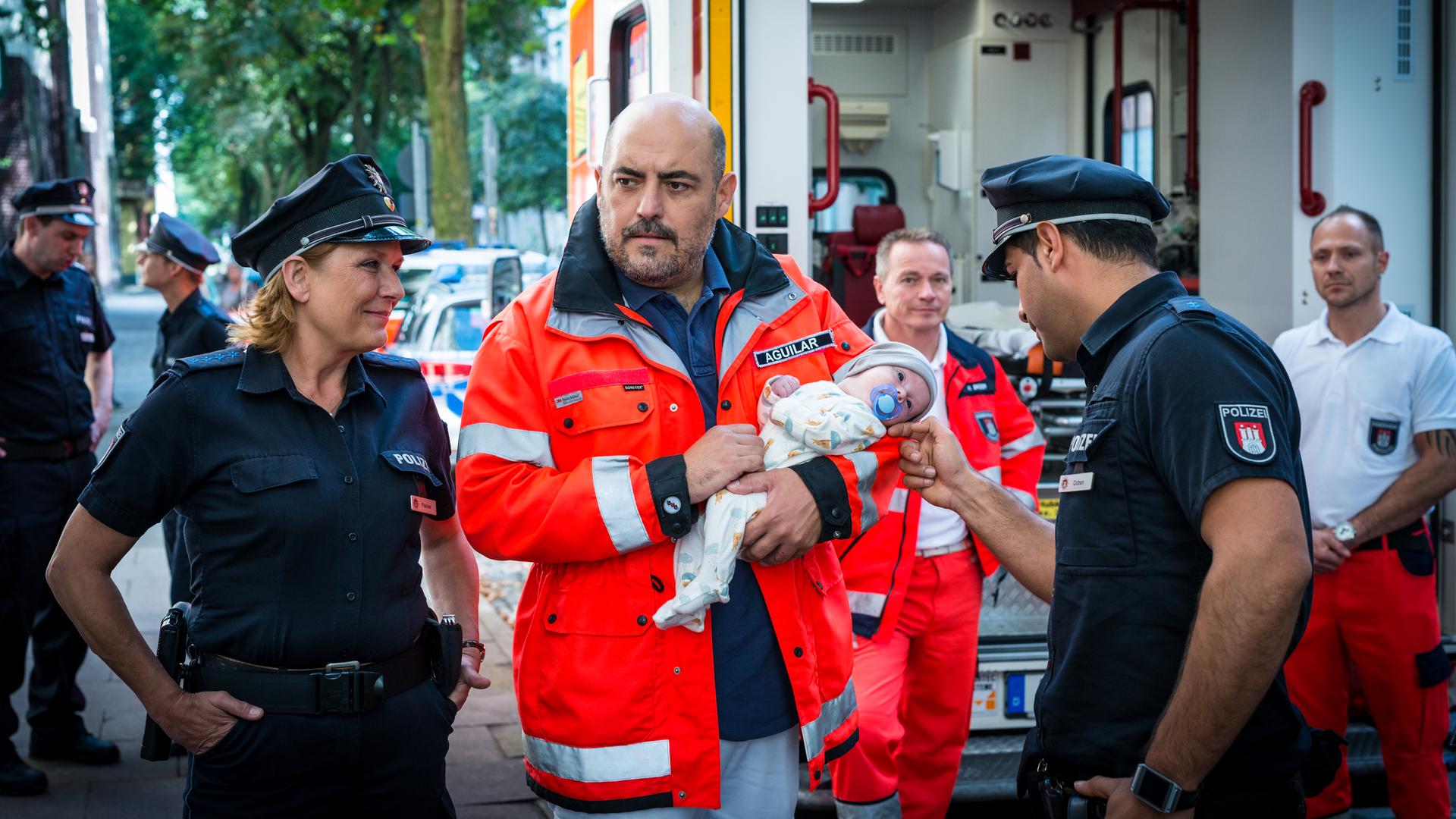 "Notruf Hafenkante - Abnabelung": Zwischen den Polizisten Claudia Fischer (Janette Rauch) und Tarik Coban (Serhat Cokgezen) steht der Arzt Dr. Aguilar (Gabriel Munoz Munoz). Er hält einen Säugling im Arm. Tarik streichelt das Gesicht des Säuglings, Claudia hat die Hände in die Hosentaschen gesteckt und schaut grinsend zu Tarik und dem Baby. Im Hintergrund der Notarztwagen und zwei Sanitäter (Komparsen).