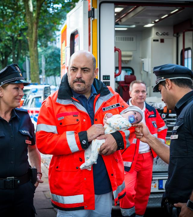 "Notruf Hafenkante - Abnabelung": Zwischen den Polizisten Claudia Fischer (Janette Rauch) und Tarik Coban (Serhat Cokgezen) steht der Arzt Dr. Aguilar (Gabriel Munoz Munoz). Er hält einen Säugling im Arm. Tarik streichelt das Gesicht des Säuglings, Claudia hat die Hände in die Hosentaschen gesteckt und schaut grinsend zu Tarik und dem Baby. Im Hintergrund der Notarztwagen und zwei Sanitäter (Komparsen).