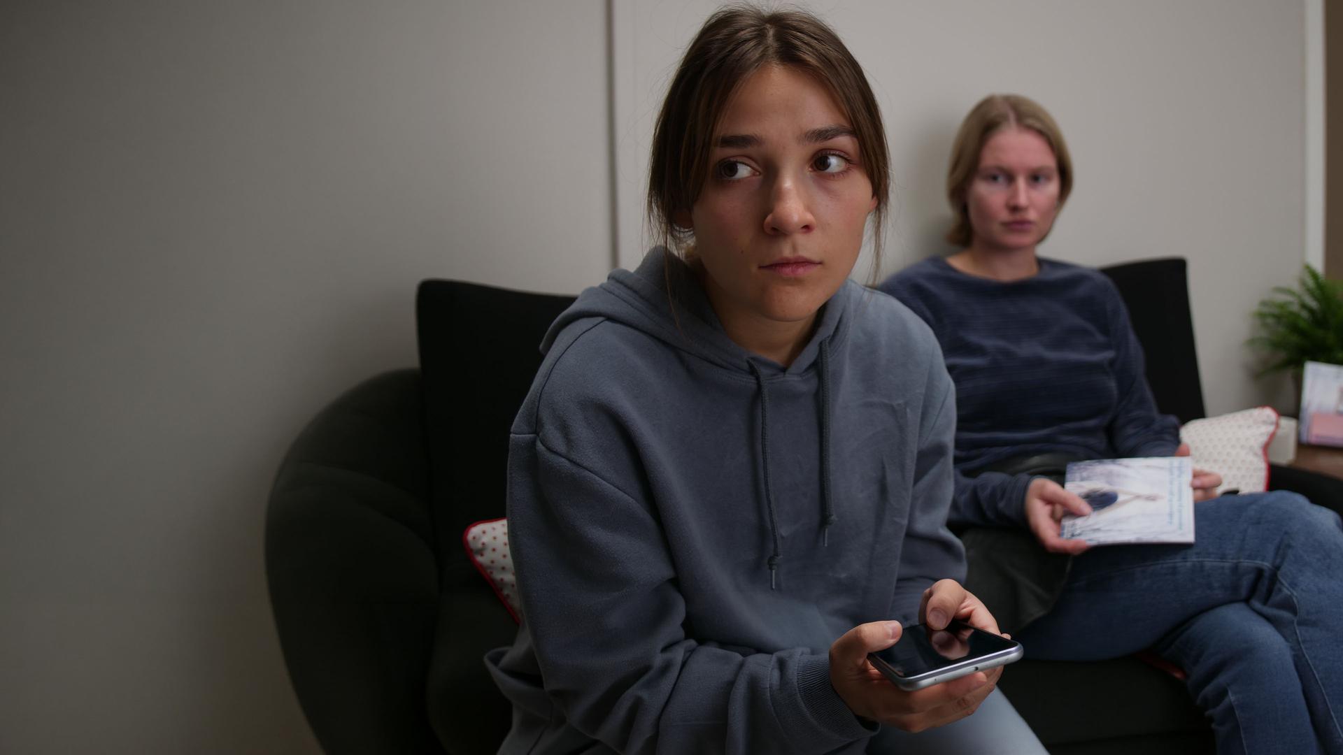 "About Saturday - Hätte Nein sagen können": Lotte (Hanna Heider Hov) sitzt hinter Klara (Darin Hagi) auf einem Sofa und schaut sie an, während Klara geistesabwesend in die Ferne blickt.