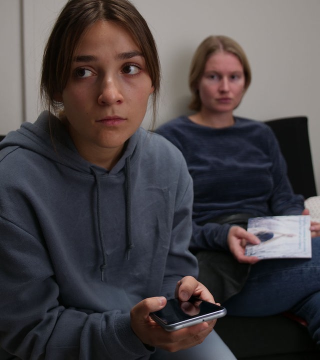 "About Saturday - Hätte Nein sagen können": Lotte (Hanna Heider Hov) sitzt hinter Klara (Darin Hagi) auf einem Sofa und schaut sie an, während Klara geistesabwesend in die Ferne blickt.