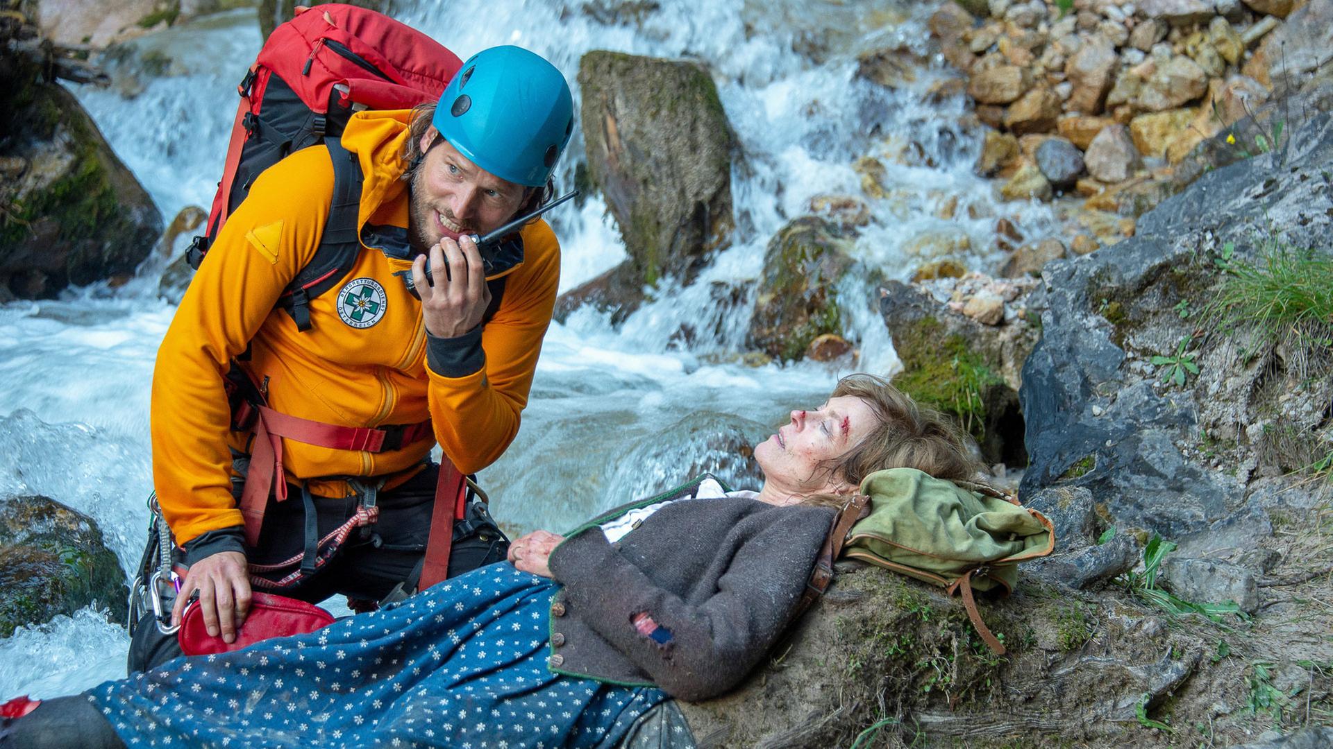 "Die Bergretter: Abschied für immer": Am Rand eines Wildwasserflusses liegt schwer verletzt Lotta Steyer (Babett Arens). Der Bergretter Markus Kofler (Sebastian Ströbel) kniet neben ihr und spricht in sein Funkgerät.