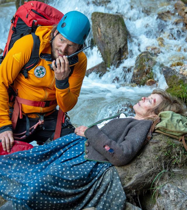 "Die Bergretter: Abschied für immer": Am Rand eines Wildwasserflusses liegt schwer verletzt Lotta Steyer (Babett Arens). Der Bergretter Markus Kofler (Sebastian Ströbel) kniet neben ihr und spricht in sein Funkgerät.