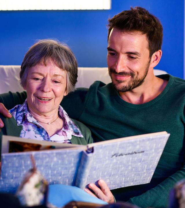 "Bettys Diagnose - Abschiedsschmerz": Dr. Frank Stern (Max Alberti) sitzt mit Oma Ilse (Monika Lennartz) im Krankenbett. Sie schauen sich lächelnd alte Fotos an.