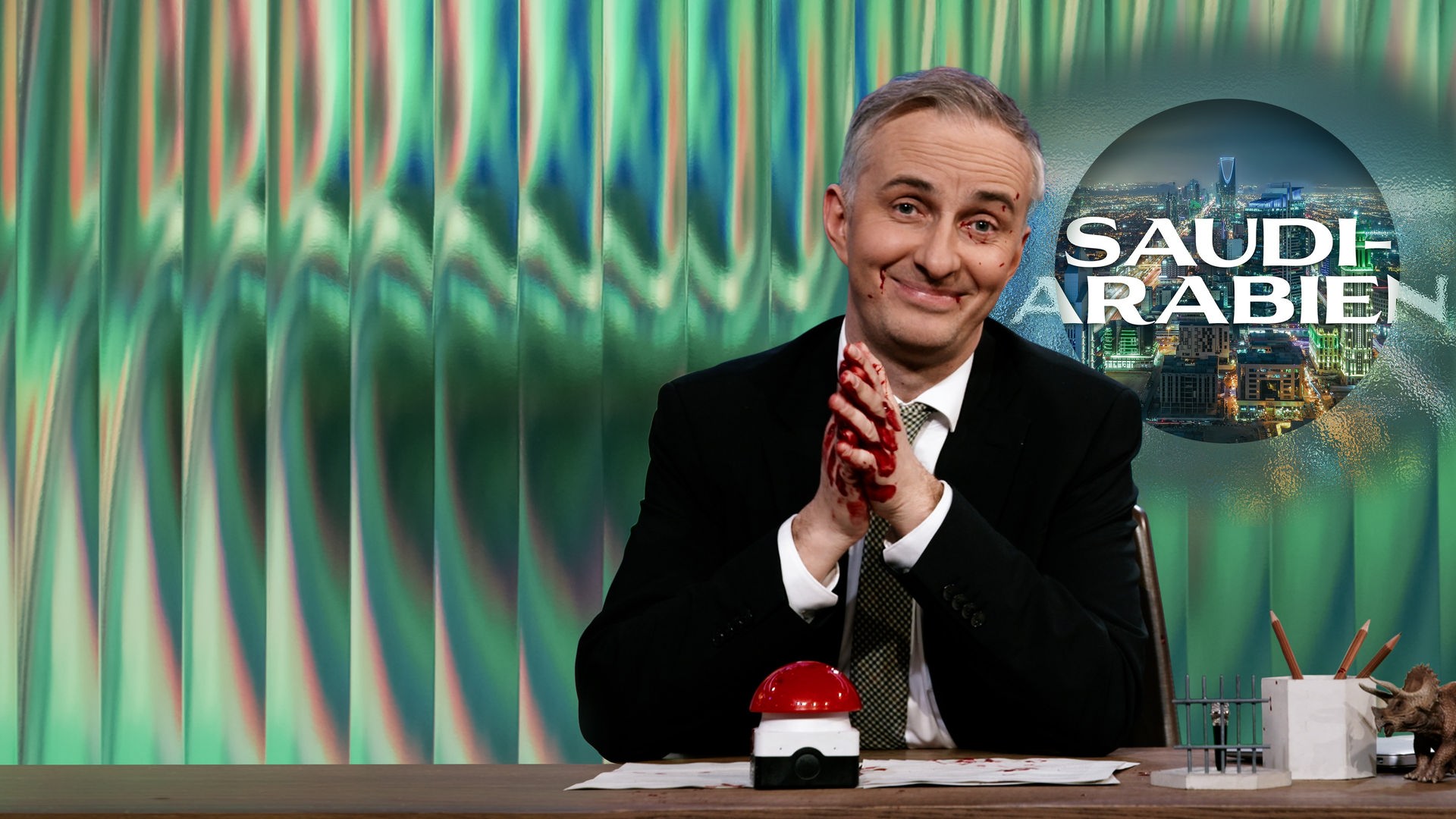 Jan Böhmermann sitzt am Schreibtisch. Er hat Blut an den Händen und im Gesicht. Rechts von ihm ist eine nächtliche, moderne Skyline zu sehen, darüber liegt der Schriftzug "Saudi-Arabien".