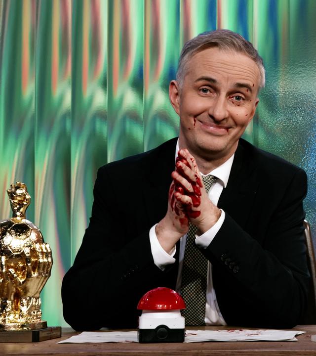 Jan Böhmermann sitzt am Schreibtisch. Er hat Blut an den Händen und im Gesicht. Rechts von ihm ist eine nächtliche, moderne Skyline zu sehen, darüber liegt der Schriftzug "Saudi-Arabien".